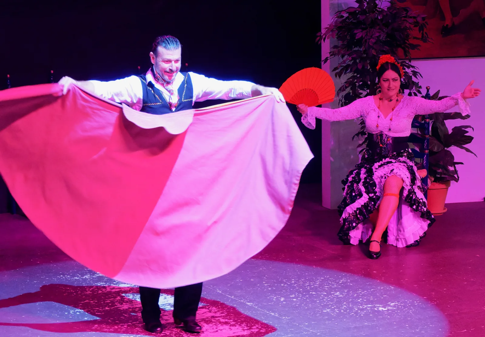 A male dancer in a waistcoat spreads a large red and white cape wide on a spotlit stage while a female dancer in a black polka-dot dress sits with a red fan raised, both caught mid-performance